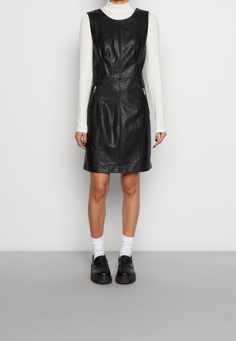 Schwarzes ledernes ärmelloses Kleid mit taillierter Silhouette, Nahtdetails an der Vorderseite und seitlichen Reißverschluss-Taschen, kombiniert mit einem weißen Langarmoberteil und schwarzen Schuhen.