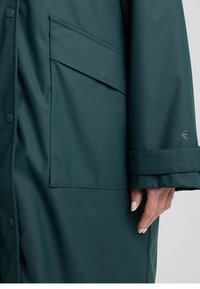 Manteau imperméable vert foncé avec une grande poche frontale, fermetures à boutons et manches retroussées. Texture lisse, design minimaliste et détail du logo.