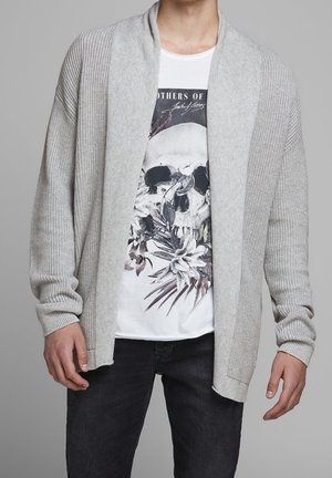 Mann trägt hellgrauen offenen Cardigan über weißem T-Shirt mit Schädel- und Blumendesign, kombiniert mit dunklen Jeans, stehend vor grauem Hintergrund.