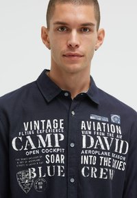 Marineblaue Button-up-Hemd mit weißem Drucktext und Grafiken, ausgestattet mit einem klassischen Kragen und strukturiertem Stoff.