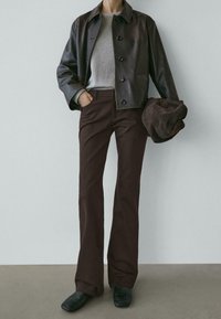 Hnědá kožená bunda s černými knoflíky, šedý svetr a tmavě hnědé rozšířené kalhoty. Vzhled doplňují černé boty a v ruce hnědý látkový klobouk.