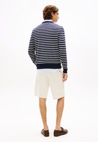Πουλόβερ με ναυτικό μπλε και λευκές ρίγες, μείγμα βαμβακιού, μονόχρωμη ναυτική μπλε ταινία στο τελείωμα· συνδυασμένο με μπεζ σορτς· το μοντέλο στέκεται με την πλάτη στην κάμερα, καφέ παπούτσια.