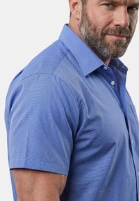Hombre con barba, vestido con una camisa azul de manga corta abotonada, mirando ligeramente de lado hacia la cámara.