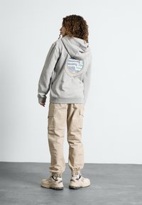 Sweatshirt cinza com um logótipo nas costas, combinado com calças cargo bege e ténis claros, apresentando punhos elásticos e múltiplos bolsos.