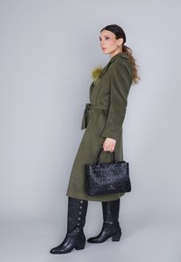 Cappotto verde oliva con vita legata, caratterizzato da un accent yellow testurizzato; abbinato a una borsa nera goffrata e stivali neri sopra il ginocchio.