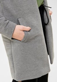 Grau gerippter Cardigan mit strukturierter Oberfläche, ausgestattet mit einer Fronttasche und einem offenen Design, geschlossen mit einem schwarzen Kordelzug.