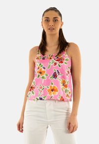 Top de tirantes floral en tela de un brillante color rosa, con un escote con volantes, que presenta coloridos patrones de flores en naranja, púrpura y verde. Combinado con pantalones blancos.