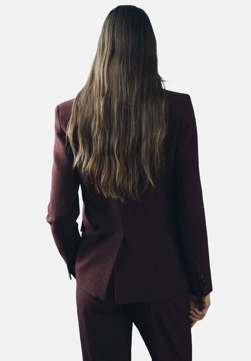 Personne aux longs cheveux bruns portant un costume rayé bordeaux foncé, debout dos au spectateur sur un fond uni.