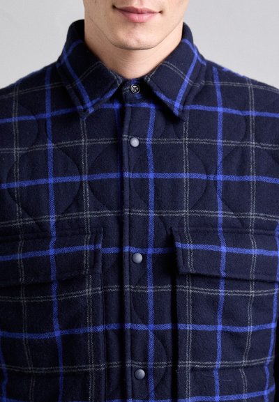 Navy plaid shirt with blue and gray lines, quilted texture, two front pockets, and snap button closure at the collar.