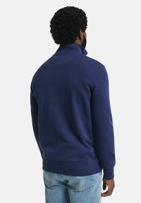 Uomo con capelli ricci che indossa un pullover blu navy con colletto alto e jeans azzurri, girato di spalle su uno sfondo bianco.