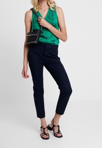 Top senza maniche in satin verde, pantaloni su misura blu scuro, borsa nera decorata e scarpe con tacco nere a strappo. Tessuti lisci, design a gamba affusolata.