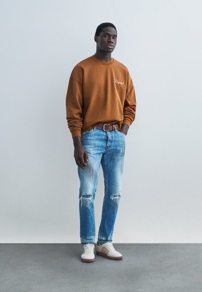 Άνδρας που στέκεται φορώντας καφέ φούτερ, ανοιχτό μπλε σκισμένο τζιν, καφέ ζώνη και λευκά αθλητικά παπούτσια μπροστά σε απλό ανοιχτό φόντο.