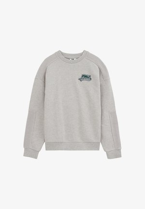 Sweat-shirt gris clair à manches longues avec poignets et épaules côtelés, avec un petit patch vert et blanc sur la poitrine gauche.