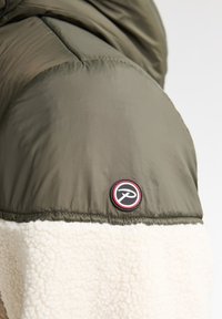 Groene geïsoleerde jas met een witte sherpa onderkant. Heeft een cirkelvormig zwart en rood logo op de schouder. Gladde en gestructureerde materialen.