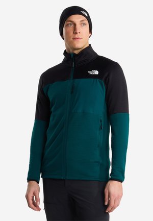 Mand iført sort og turkis lynlåstrøje og sort beanie med The North Face-logoer, stående mod en hvid baggrund.