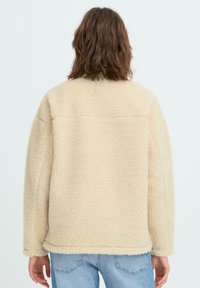 Bézs színű, bolyhos polár dzseki laza szabással, kontrasztos felülettel és varrásos részletekkel. Egyenes szegéllyel és leejtett vállrészlettel rendelkezik.