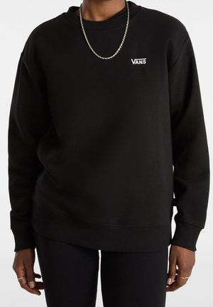 Personne portant un sweat-shirt Vans noir avec un petit logo blanc sur la poitrine et un collier chaîne argenté, debout devant un fond uni.