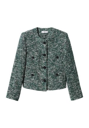 Model draagt een groene gestructureerde jas met knopen, een zwarte minirok en kniehoge zwarte leren laarzen tegen een effen achtergrond.