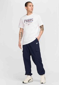 Hvid t-shirt med "PARIS SAINT-GERMAIN" i rød og blå, parret med marineblå cargo-bukser og cremede sneakers med et tekstureret design.