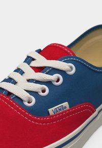 Baskets Vans rouges et bleues avec des lacets blancs et des coutures, dotées d'une semelle en caoutchouc et d'une étiquette avec le logo Vans sur le côté.