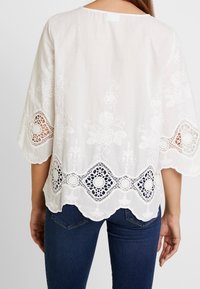 Blusa bianca con ricami floreali, orlo scallop e dettagli in pizzo sulle maniche. Abbinata a jeans blu, che mostrano una vestibilità comoda.