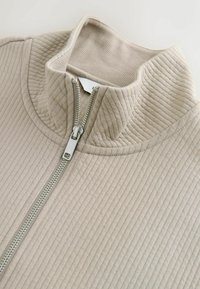 Hellbeige geripptes Stoffkleidungsstück mit einem silbernen Reißverschluss und einem Stehkragen, in Nahaufnahme gezeigt.