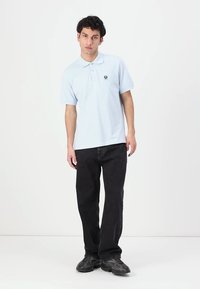 Ανοιχτό μπλε πόλο πουκάμισο με κοντά μανίκια και γιακά, με μαύρο λογότυπο στο στήθος. Συνδυασμένο με μαύρα τζιν κατευθείαν και σκούρα παπούτσια.