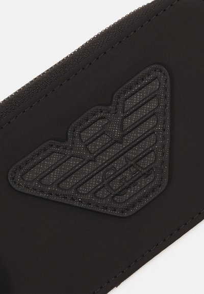 Portafoglio in tessuto nero con una toppa logo nera testurizzata a forma di ali. Le cuciture sono visibili lungo i bordi.