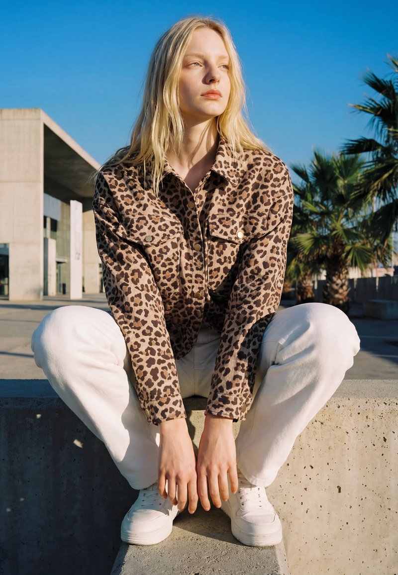 Joven mujer con cabello rubio largo que lleva una chaqueta con estampado de leopardo, pantalones blancos y zapatillas, agachada en un borde de hormigón al aire libre.