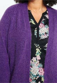 Cardigan viola lavorato a maglia con una texture morbida e pelosa; presenta la parte anteriore aperta e maniche larghe. Indossato sopra una camicetta nera con motivo floreale.