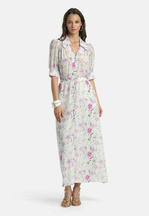 Vrouw draagt een witte maxi-jurk met bloemenprint, pofmouwen, knopen aan de voorkant, een tailleband, gouden sandalen, oorringen en witte armbanden.