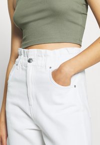 Bílé džínové kalhoty s nařaseným pasem, předními kapsami a stříbrnými kovovými doplňky. Spárovány s olivově zeleným pruhovaným crop topem.