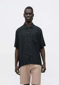 Camisa preta de manga curta com botões, um único bolso no peito, feita de tecido leve, combinada com calções texturados bege claro.