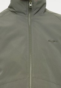 Chaqueta gris resistente al agua con cuello alto, cierre de cremallera completo y detalle sutil de logotipo. Presenta una textura suave y costuras precisas.
