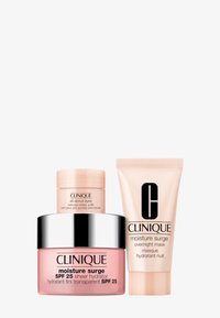 Le set de soins de la peau Clinique comprend un pot rose de Moisture Surge, un contenant noir et argenté, et un tube beige, tous avec des étiquettes détaillées.