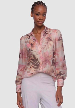 Donna con capelli intrecciati che indossa una camicetta trasparente rosa a fiori infilata in pantaloni grigi chiaro a vita alta, in piedi davanti a uno sfondo semplice.