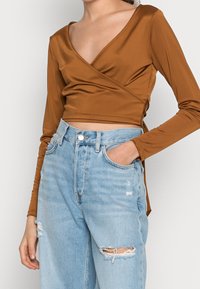 Top avvolgente marrone a maniche lunghe realizzato in tessuto liscio, con un design a scollo a V profondo. Abbinato a jeans in denim azzurri chiari e strappati.