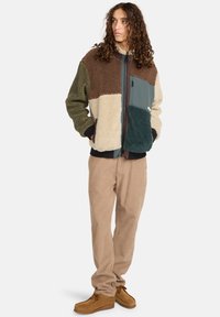 Veste en polaire avec un design en patchwork beige, vert olive, marron et teal, fermeture éclair à l'avant et deux poches ; assortie avec un pantalon beige clair.