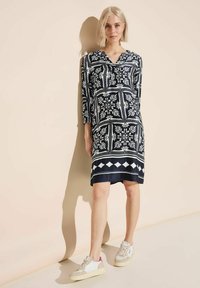 Navyblaues Kleid mit weißem Blumenmuster, V-Ausschnitt, Dreiviertelärmel und knie-langer Saum. Kombiniert mit weißen und beigen Turnschuhen.