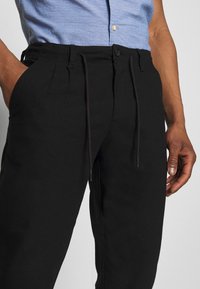 Pantaloni neri con vita regolabile, caratterizzati da una texture liscia e tasche laterali. Abbinati a una camicia azzurro chiaro con motivo e bottoni.