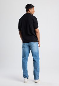Black polo shirt with short sleeves, ribbed collar, paired with light blue denim jeans. Model wears white sneakers, showcasing a casual fit.