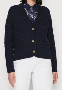 Marinblå stickad cardigan med strukturerat mönster, V-ringning, ribbad nederdel och muddar, försedda med gyllene knappar fram.