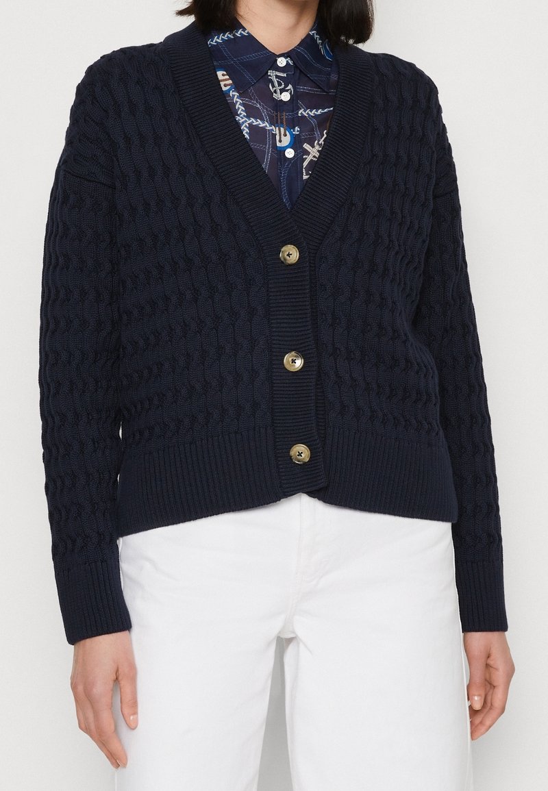 Marinblå stickad cardigan med strukturerat mönster, V-ringning, ribbad nederdel och muddar, försedda med gyllene knappar fram.
