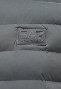 Sivi puffer jakna od materijala s kvadratnim uzorkom. Ima teksturiranu oznaku s natpisom "EA7 EMPORIO ARMANI" koja je istaknuta.