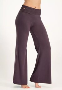 Person som bär mörklila vida byxor med elastisk midja, står barbent på en ljus bakgrund.