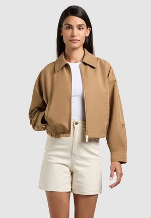 Donna con capelli lunghi e scuri che indossa una giacca marrone chiaro con zip, una maglietta bianca e pantaloncini crema a vita alta su uno sfondo chiaro e uniforme.