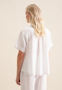 Camicia bianca in lino a maniche corte con una vestibilità comoda, dotata di una piega sul retro e una texture sottile, abbinata a pantaloni chiari coordinati.