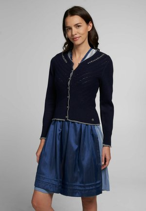 Cardigan en maille bleu marine avec un col en V, des bordures grises, des coutures décoratives et des boutons argentés, associé à une jupe en satin bleu avec des plis.