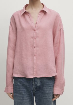 Personne portant une chemise rose ample à manches longues, boutonnée, rentrée sur un jean en denim bleu.