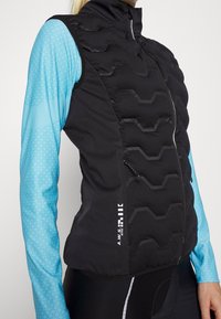 Person som bär en svart vadderad väst över en långärmad blå mönstrad träningsskjorta och svarta byxor med vit sidodetalj.